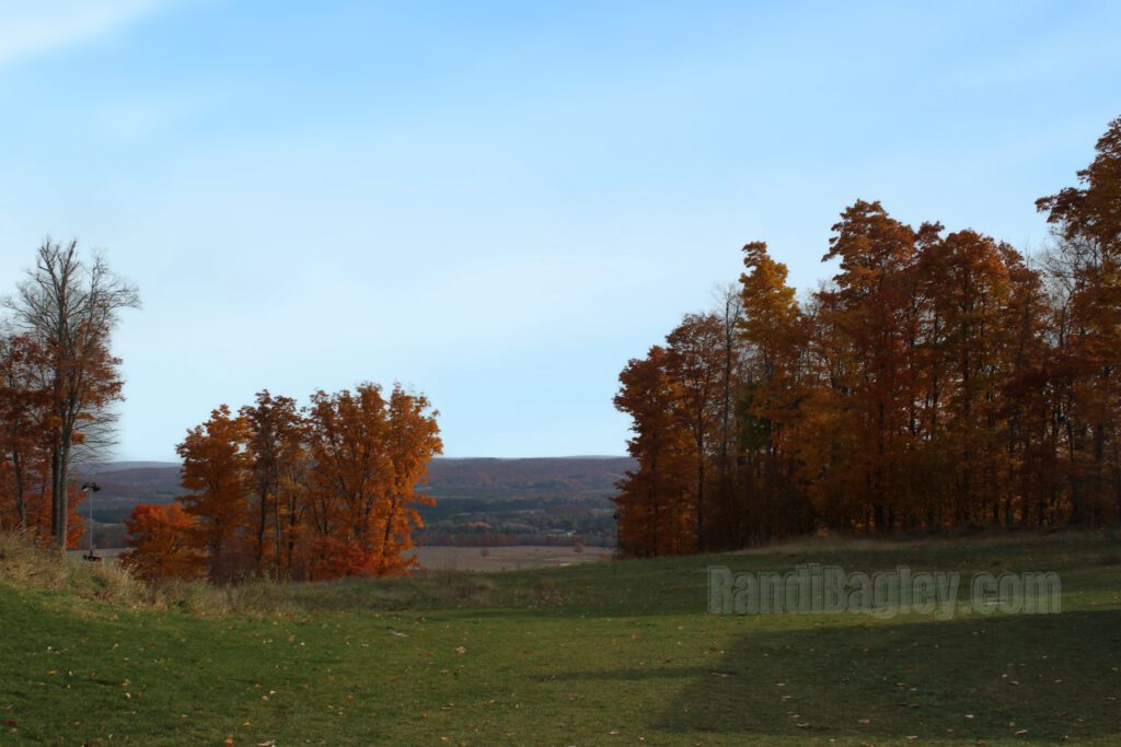 Open green space with fall-colored trees and rolling hills in the distance under a blue sky.